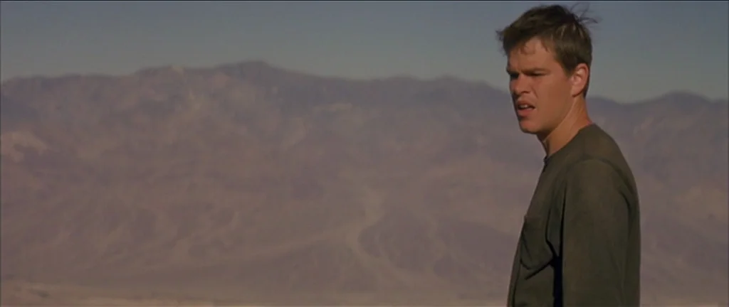 Gerry (2002) movie still: medium — A vast, dry desert mountain range stretches across the horizon under a clear sky. A young man wearing a green…