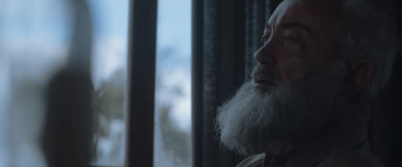 My Neighbor Adolf (2022) movie still: closeup — A dimly lit interior space features a man sitting beside a glass window. He looks toward the light…