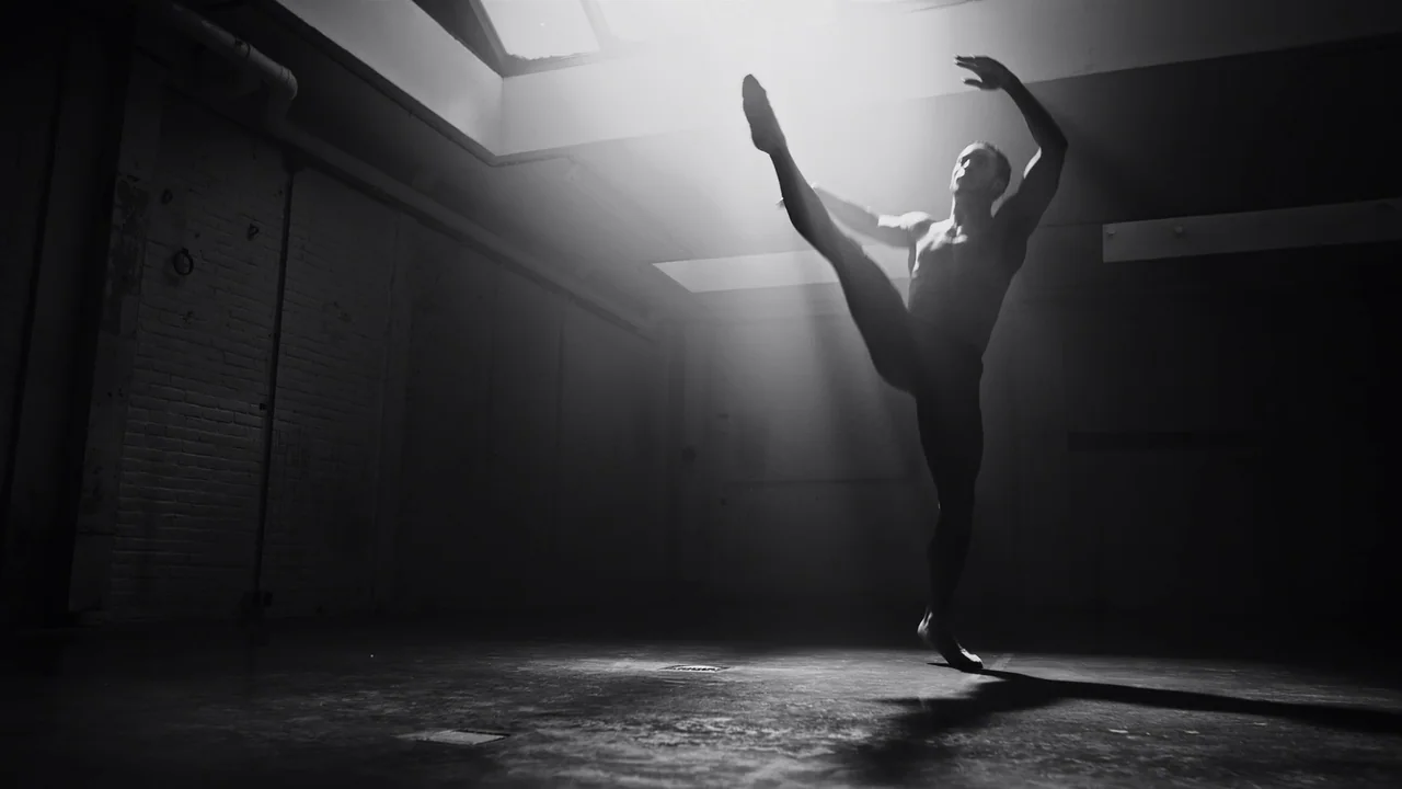 Kokomo City (2023) movie still: fullbody — An industrial-style interior with brick walls and large overhead windows. A male dancer stands on one leg…