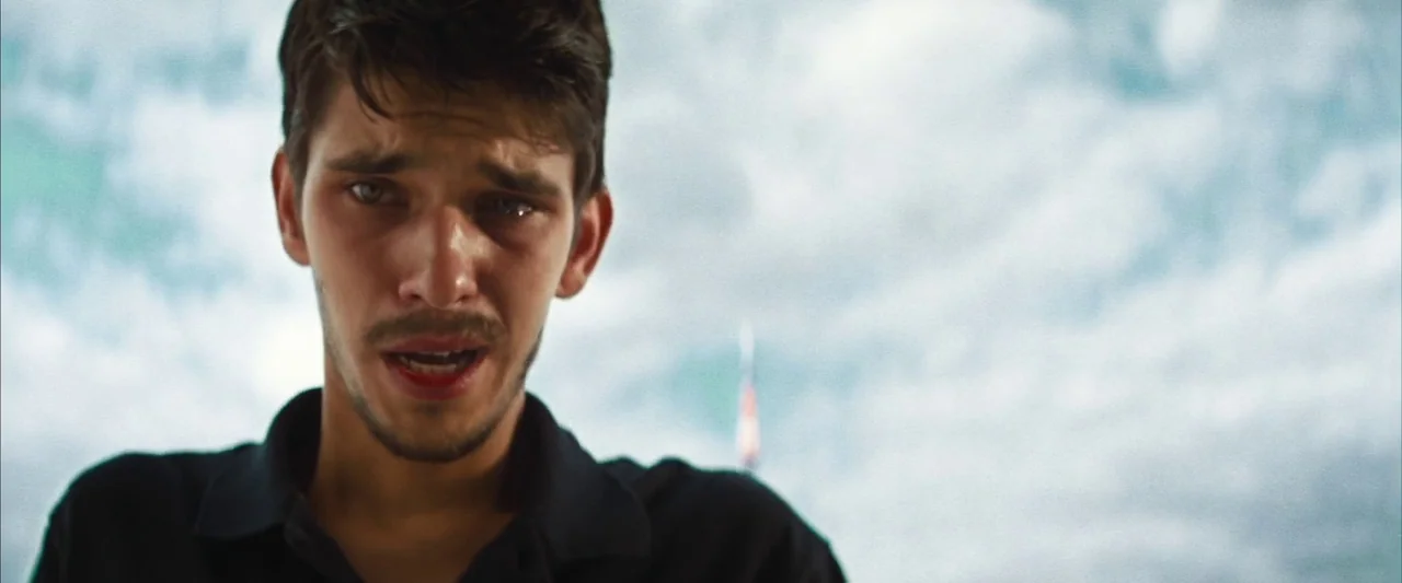 Layer Cake (2004) movie still: closeup — The frame captures a young man outdoors against a bright, cloudy sky. A single tear falls down his cheek…