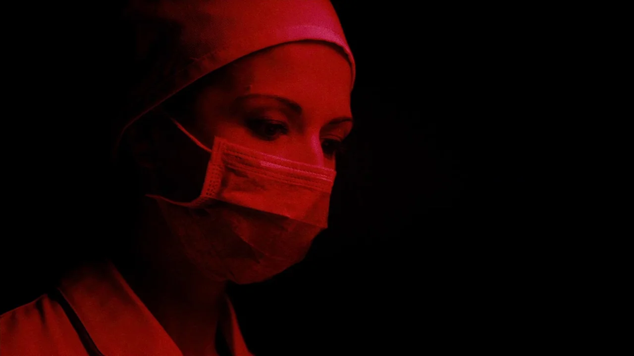 The ABCs of Death (2012) movie still: closeup — A close-up of a person in medical gear against a completely dark background. The subject is…