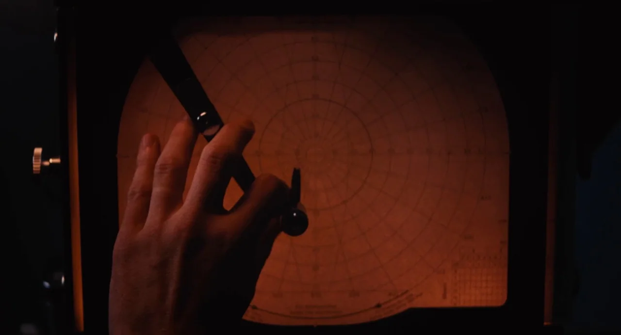 Ava (2017) movie still: closeup — An orange-glowing circular grid screen from a vintage radar console dominates the dark room. A hand holds a pair…