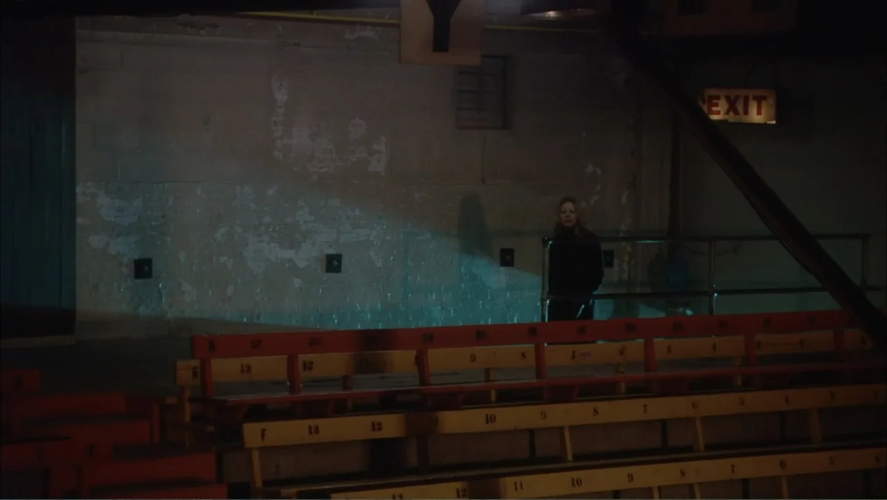 O código oculto (2025) movie still: wide — A dimly lit industrial-style seating area features numbered wooden benches and a concrete wall. A woman…