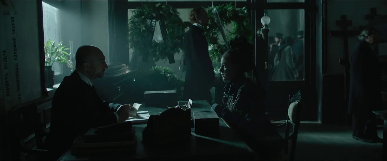 Tchaikovsky’s Wife (2022) movie still: medium — A dark funeral home interior features wooden tables and large floral funeral wreaths. A man writes…