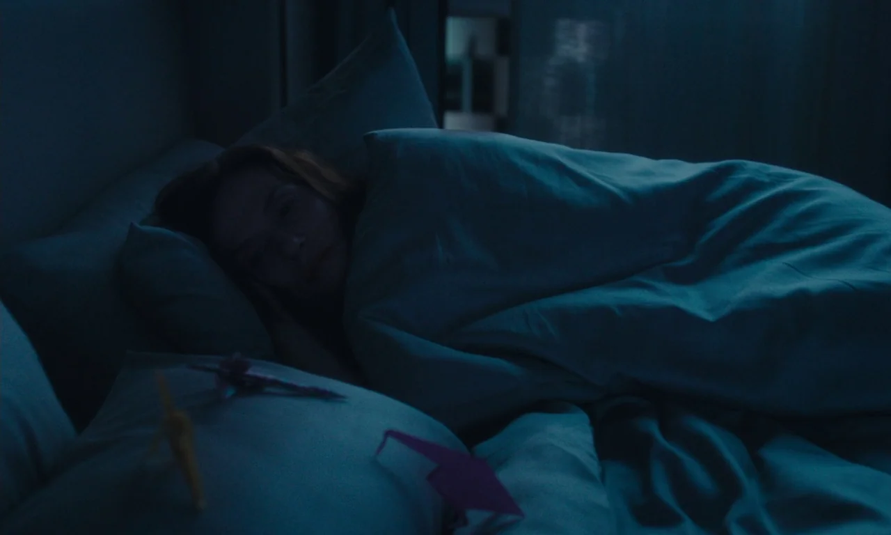 Sidonie in Japan (2023) movie still: medium — A dark, blue-toned room features a woman lying on a bed at night. Small paper cranes rest on the…