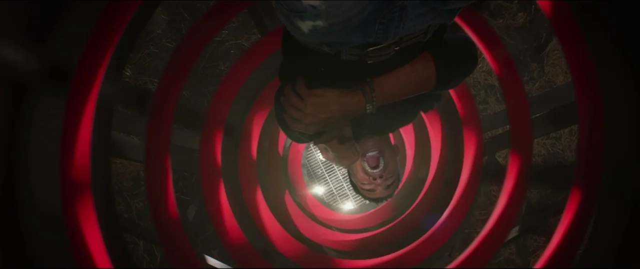 Jigsaw (2017) movie still: medium — A man slides down inside a metal red spiral structure at night. He is oriented upside down toward the camera as…