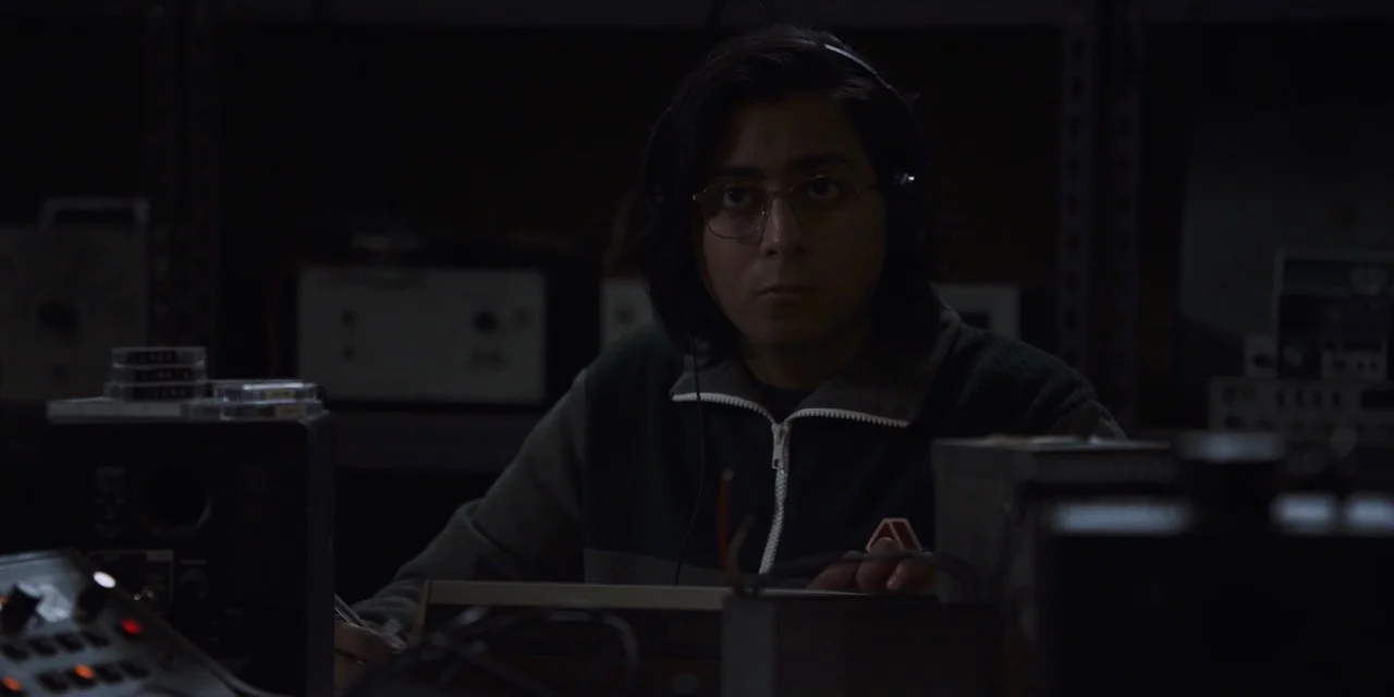 The Sound of Silence movie still: medium — A dimly lit room filled with vintage analog electronic equipment and audio recording devices. A young man…