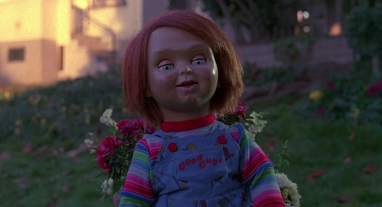 Child’s Play 2 (1990) movie still: medium — A doll sits in a grassy outdoor area near a suburban house at sunset. The figure faces the camera…