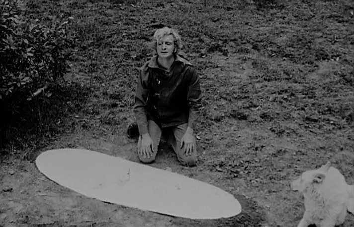 Fando y Lis (1968) movie still: medium — A man kneels on the dirt floor of a sparsely vegetated field. A large white oval object sits on the ground…
