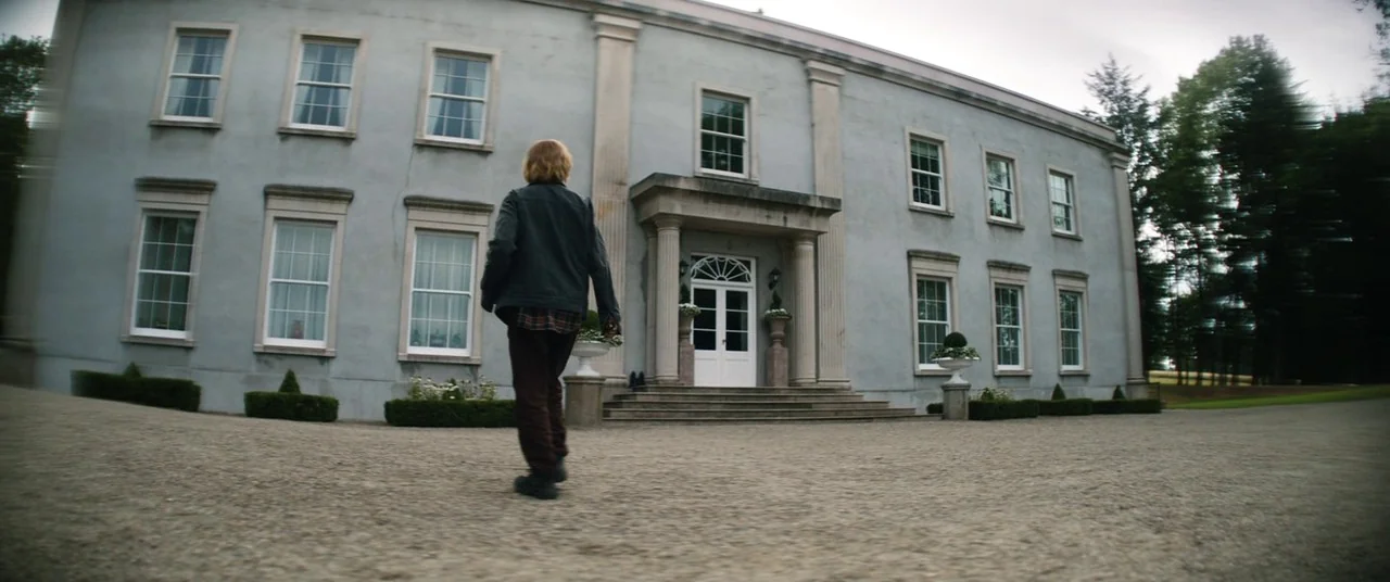 The Rhythm Section (2020) movie still: fullbody — A large grey stone manor with multiple sash windows sits behind a gravel driveway. A single person…