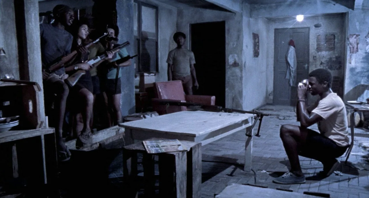 City of God (2002) movie still: wide — A dimly lit room with distressed walls and a tiled floor serves as a hideout. A man photographs a group of…