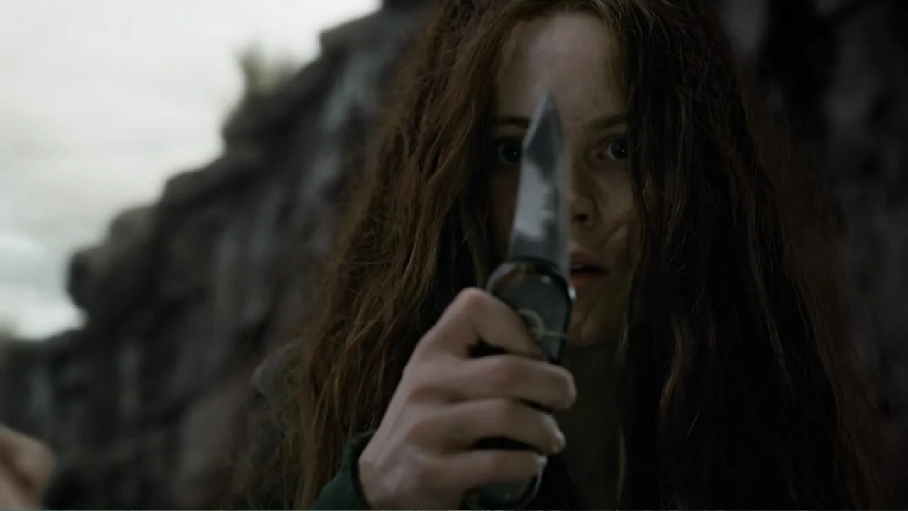 Mortal Engines (2018) movie still: closeup — A woman stands in front of a jagged grey rock wall in an outdoor setting. She holds a knife blade…
