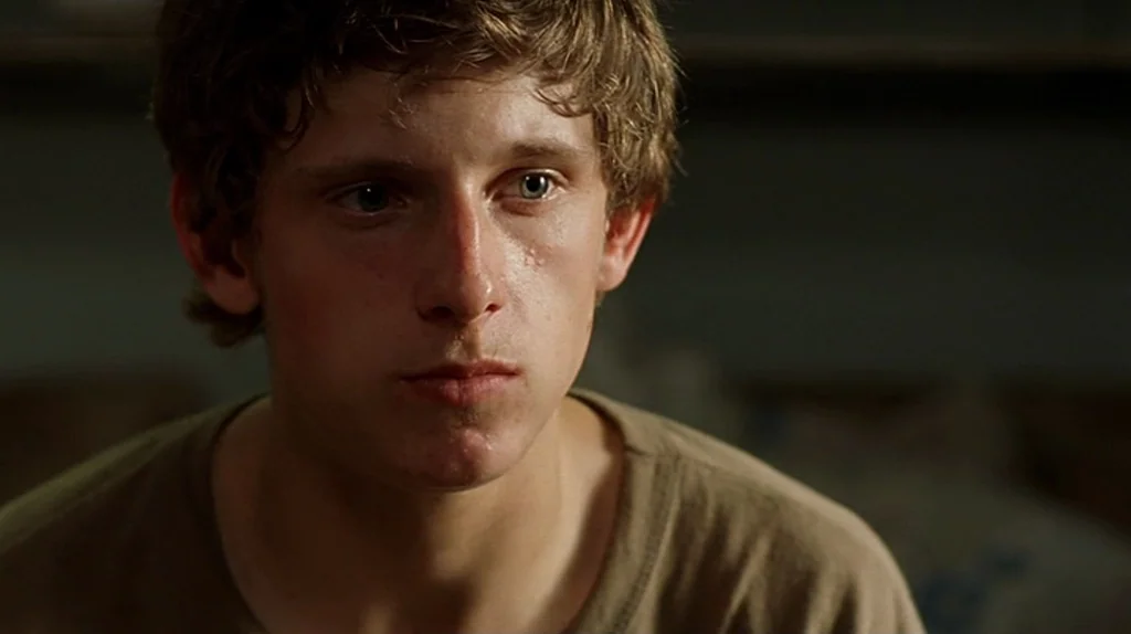 Undertow (2004) movie still: closeup — An interior space with muted colors and a dark background. A young man stares directly ahead with a still,…
