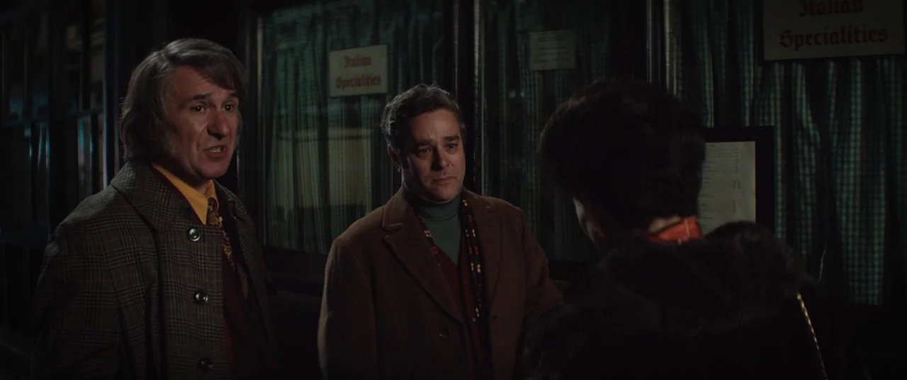 Judy (2019) movie still: medium — A night street scene outside a storefront with windows displaying Italian specialties signage. Two men stand…