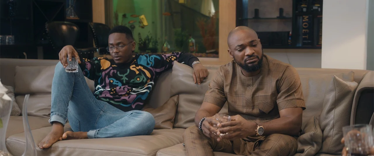 Big Love (2012) movie still: medium — A modern living room features a tan leather sectional sofa and an aquarium. Two men sit on the couch while…