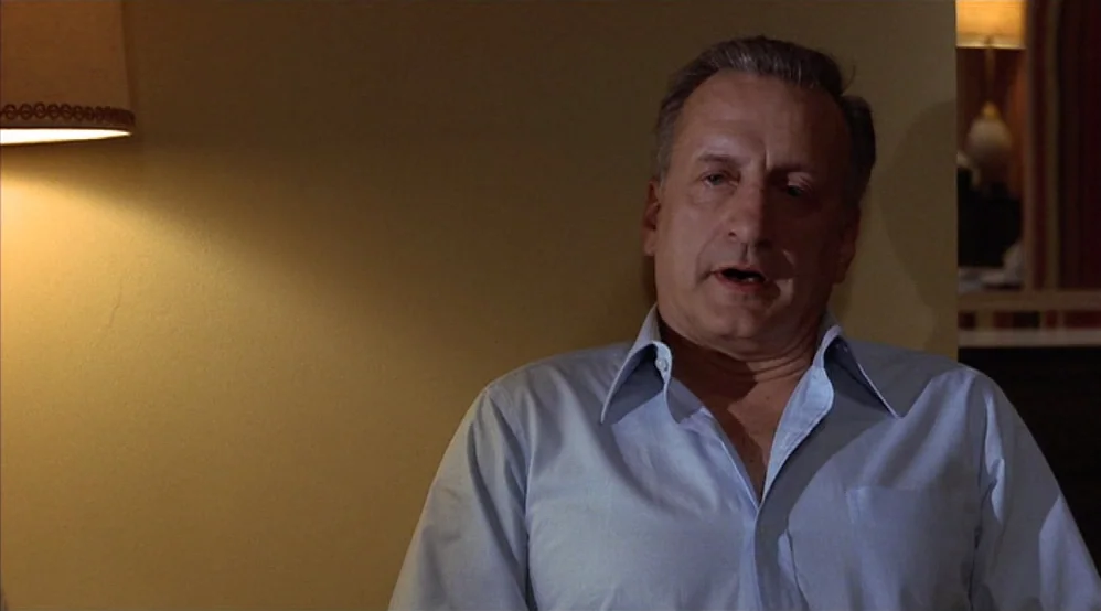 Hardcore (1979) movie still: medium — A man stands against a plain yellow wall in a dim hotel space. He is speaking while positioned near a lit wall…