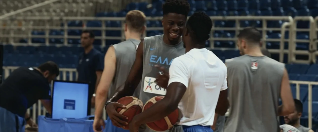 Giannis: The Marvelous Journey (2024) movie still: medium — A group of basketball players stand on an indoor court during practice. Two men in the…