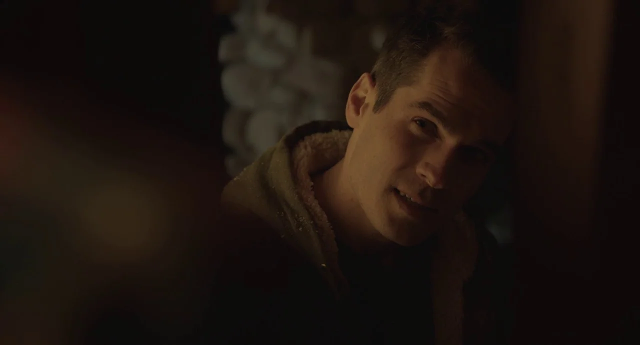 The Wolf of Snow Hollow (2020) movie still: closeup — A dimly lit room with a textured wall blurred in the background. A man wearing a hooded jacket…