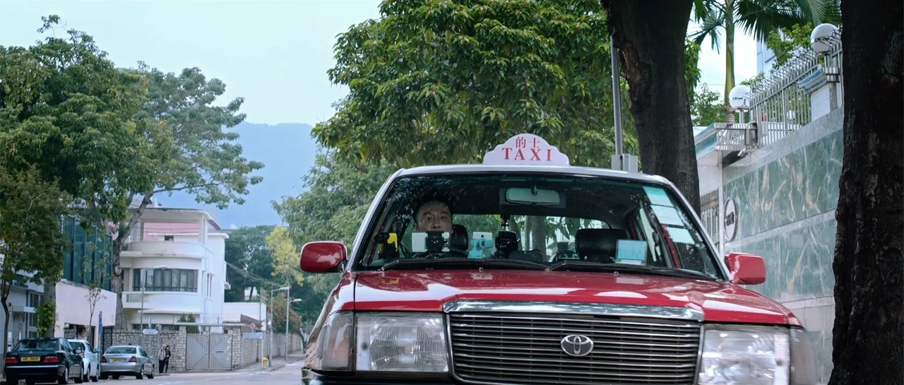 Line Walker (2016) movie still: medium — A red taxi drives down a quiet suburban street bordered by leafy trees. The middle-aged male driver looks…