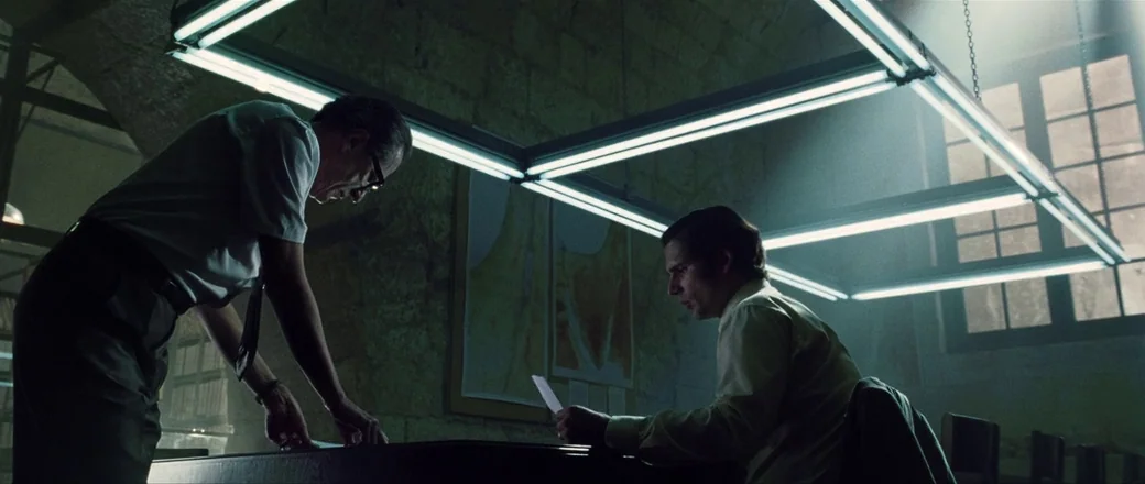 Munich (2005) movie still: medium — A stone-walled room features a large rectangular fluorescent light fixture suspended above a table. Two men…