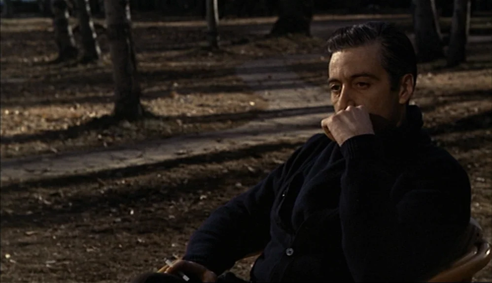 The Godfather Part II (1974) movie still: medium — A man sits in a chair in a sparse park during the autumn season. He rests his chin on his hand…