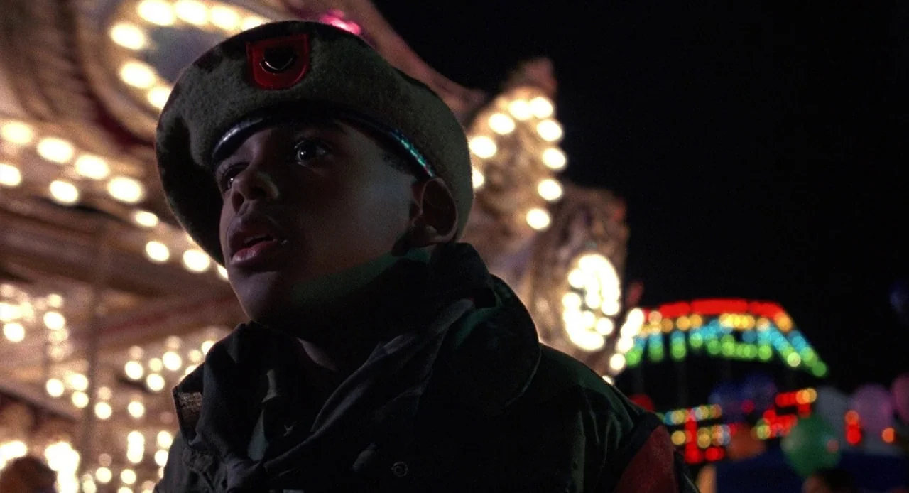 Child’s Play 3 (1991) movie still: closeup — An amusement park at night features blurred rows of bright, multi-colored light bulbs. A teenage boy…