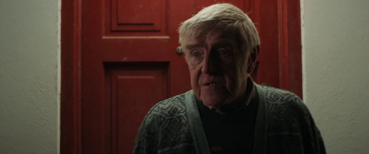 Extra Ordinary (2019) movie still: medium — An elderly man stands in a dimly lit indoor space before a solid red door. The wall beside him is…