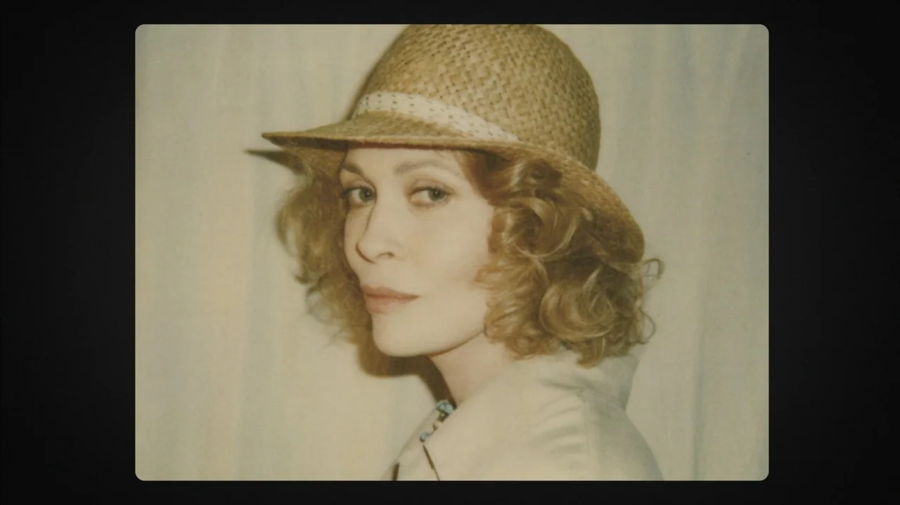 Faye (2024) movie still: closeup — A woman stands before a plain off-white cloth backdrop in an interior space. She wears a straw hat and looks…