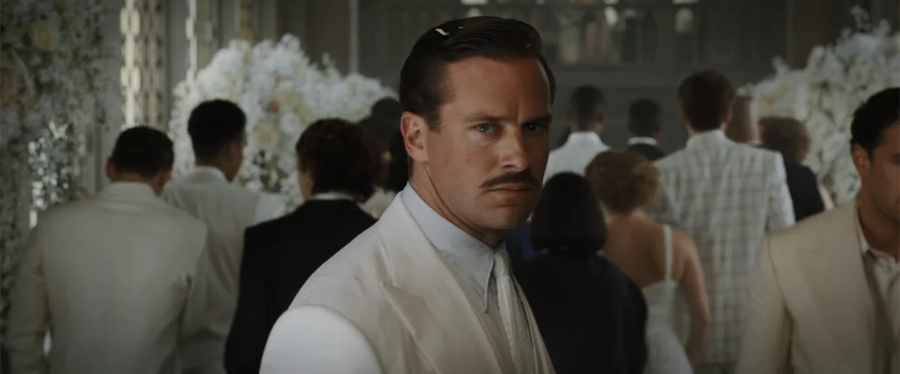 Death on the Nile (2022) movie still: medium — A crowded interior ballroom features numerous guests in formal white summer attire. A man with a dark…