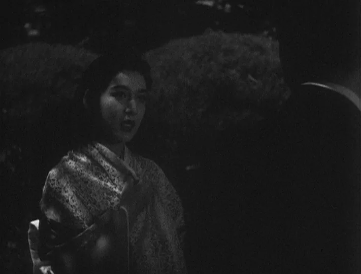 No Regrets for Our Youth (1946) movie still: medium — A woman stands in a dark outdoor setting with dense foliage in the background. She faces…