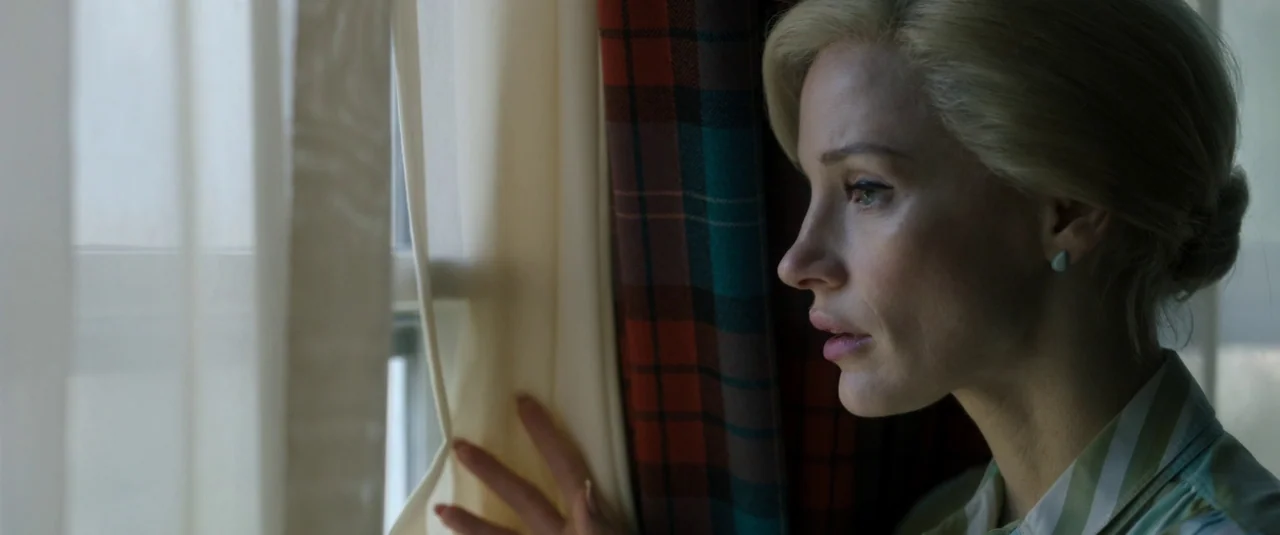 Mother’s Instinct (2024) movie still: closeup — A woman stands indoors near a window covered by sheer white curtains and plaid drapes. She looks…