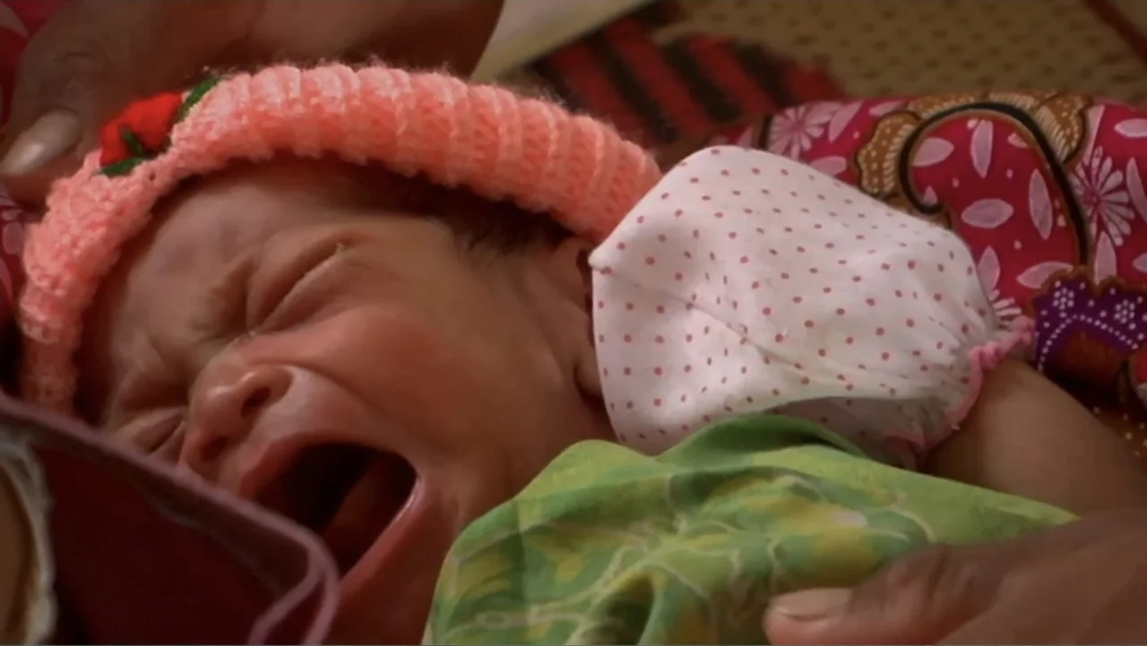 Sister (2014) movie still: closeup — A close-up view shows a newborn baby resting against a patterned fabric background. The baby wears a pink knit…