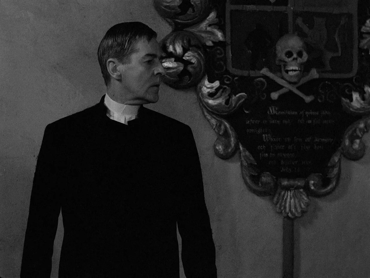 Winter Light (1963) movie still: medium — A dark stone wall in a church features an ornate carved funerary plaque. A man in clerical attire stands…