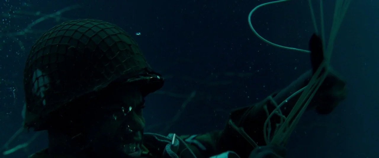 Overlord (2018) movie still: closeup — A soldier wearing a military helmet is submerged deep underwater. The man pulls on a bundle of white…