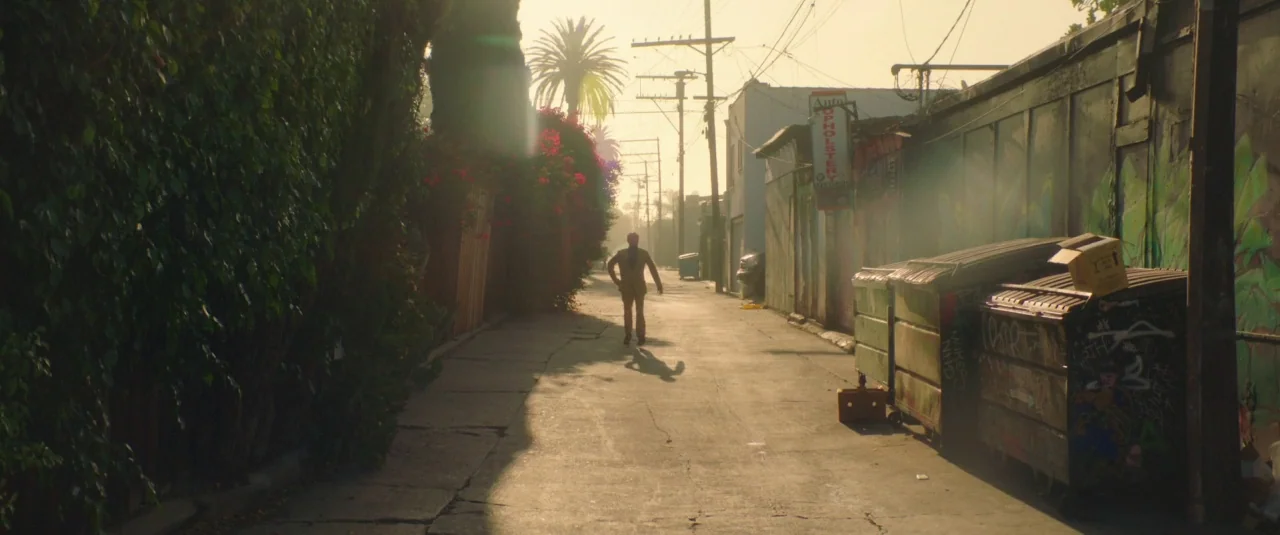 Lemon (2017) movie still: wide — An urban alleyway features a concrete path flanked by graffiti-covered walls and flowering foliage. A man in a…