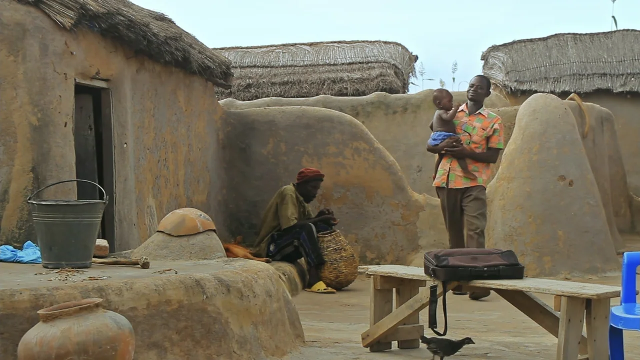 The Money Stone (2018) movie still: fullbody — A rural setting features mud-walled structures with thatched roofs and a dirt courtyard. A man…