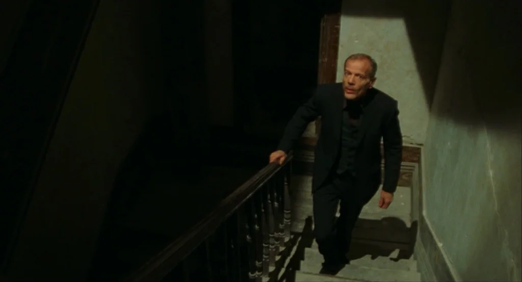 This Night (2008) movie still: medium — A dimly lit indoor stairwell features worn walls and a wooden handrail. A man in a dark suit climbs the…