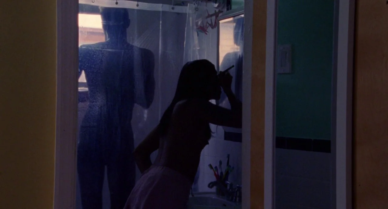 So Pretty (2019) movie still: medium — A small bathroom with a sink, mirror, and a partially obscured shower area. A woman stands by the mirror…