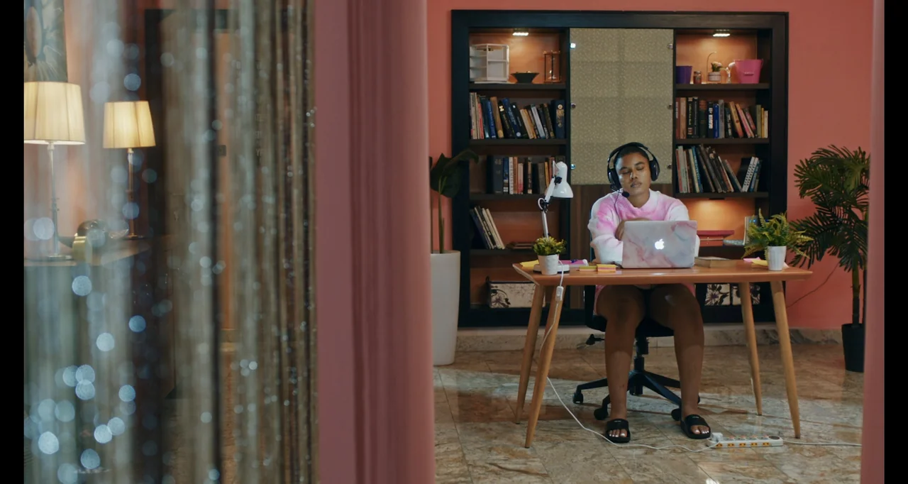 Love In A Pandemic (2023) movie still: medium — A modern home office with salmon-colored walls and a bookshelf filled with books. A young woman sits…