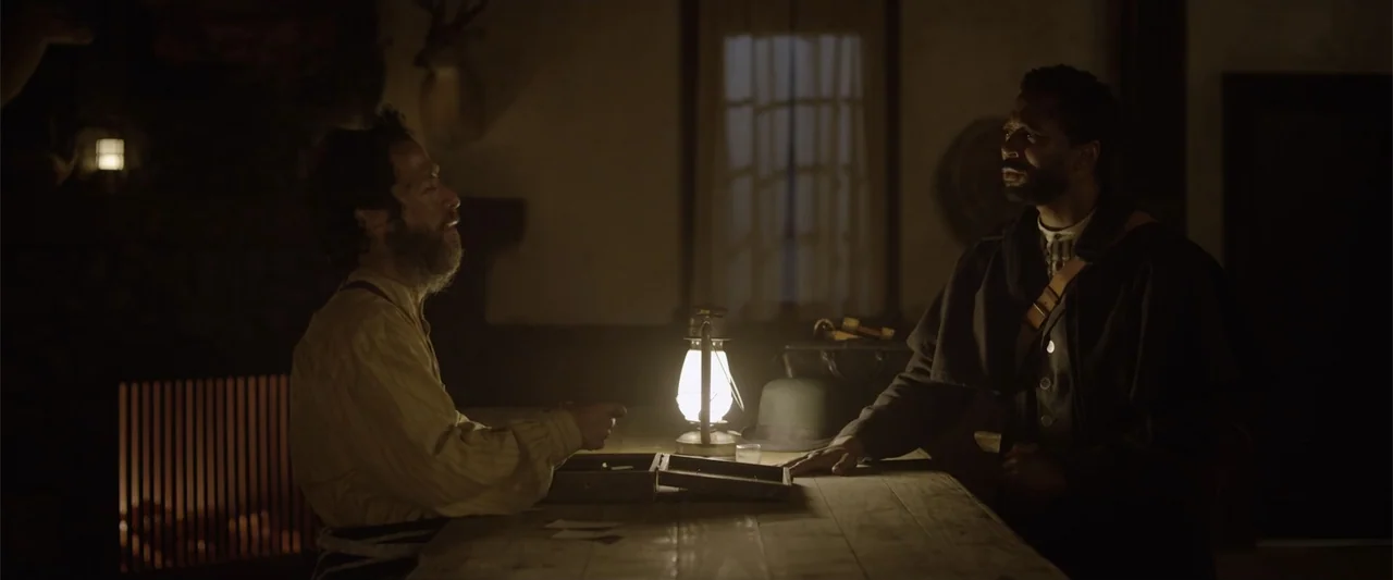 Ghosts of the Ozarks (2022) movie still: medium — A rustic wood-paneled room features a long table with a lit kerosene lantern. Two men sit at the…