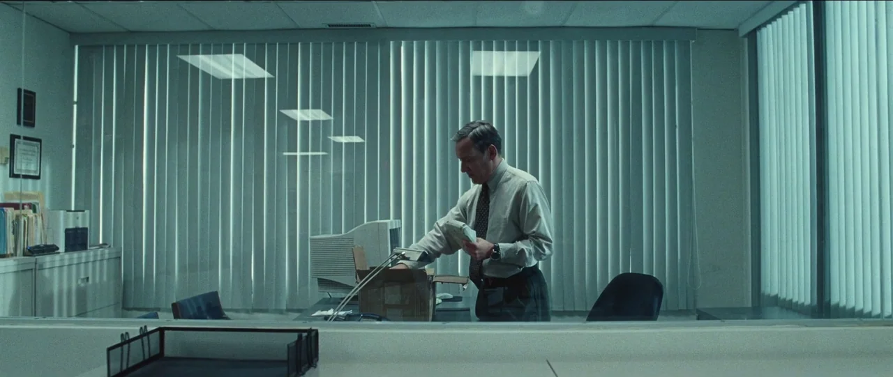 Bad Education (2019) movie still: medium — A modern 1990s cubicle is separated from the foreground by a glass partition. The man stands inside the…