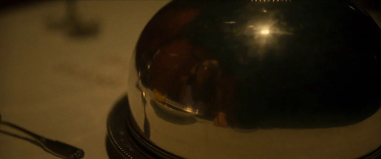 Earwig (2022) movie still: closeup — A polished silver serving cloche rests on a linen tablecloth in dim lighting. The curved surface of the metal…