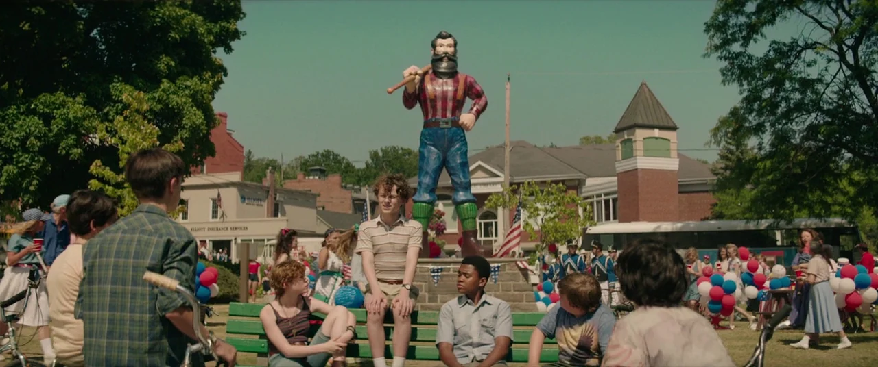 It (2017) movie still: wide — A sunny town park features a prominent statue of a lumberjack holding an axe. A group of teenagers sits on a green…