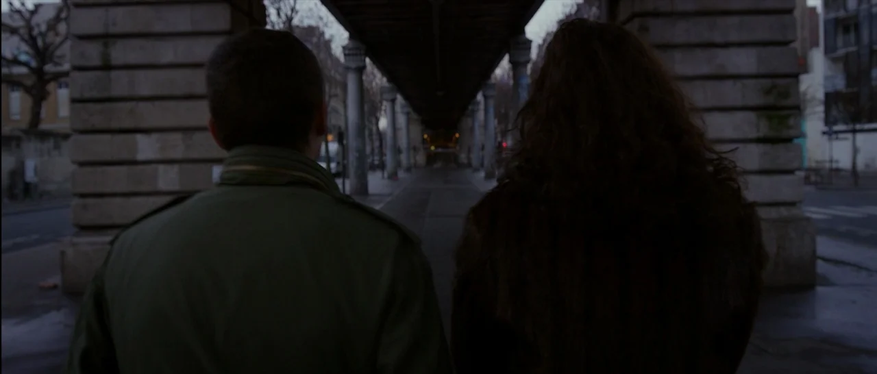 Love (2015) movie still: medium — A dark concrete underpass with repeating stone pillars stretches into the distance. Two figures walk away from the…