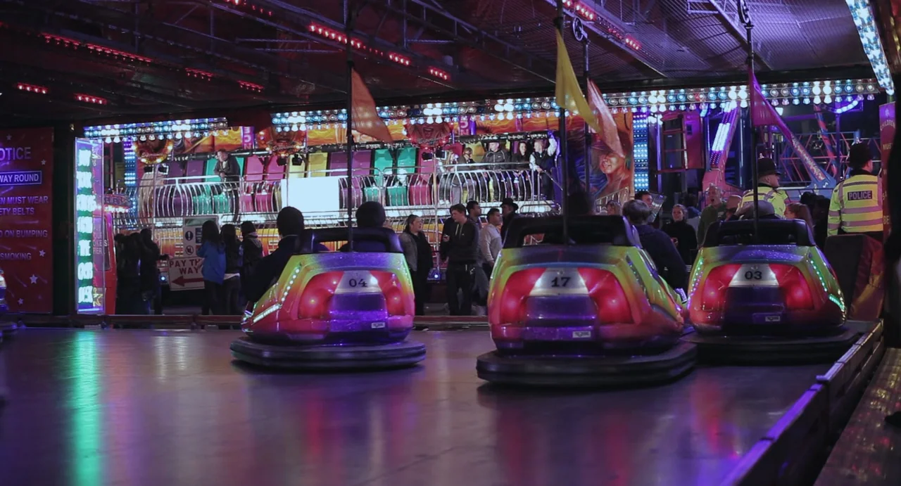 Weekend (2011) movie still: wide — A fairground bumper car attraction features numbered vehicles parked on a smooth reflective floor. People stand…
