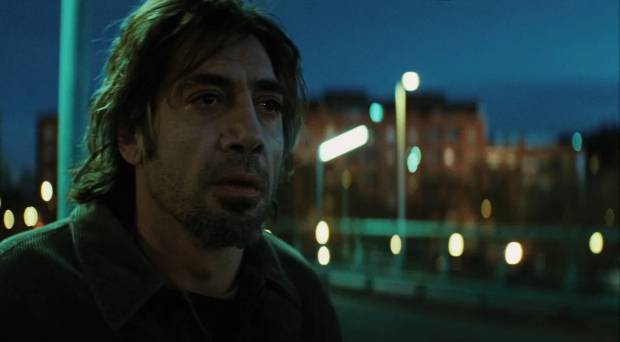 Biutiful (2010) movie still: closeup — A city street at night illuminated by distant, out-of-focus streetlights. A middle-aged man with messy hair…