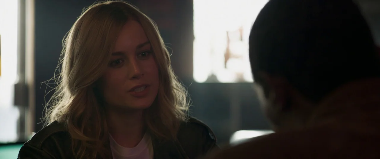Captain Marvel (2019) movie still: medium — A dark, dimly lit space with a window casting light from behind. A woman sits at a table speaking to a…