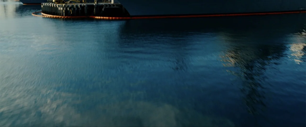 Battleship (2012) movie still: establishing — A harbor scene showing calm blue water next to a dark industrial pier structure. A bright orange…