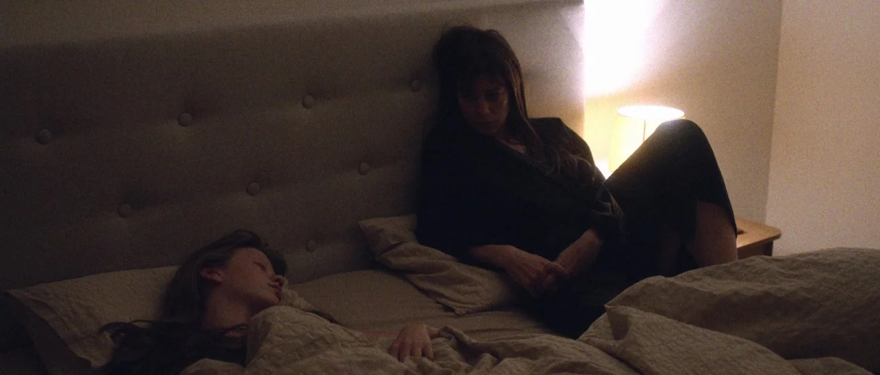 Nymphomaniac Vol 2 (2013) movie still: medium — A dimly lit room features a large bed with a button-tufted headboard. One woman sits awake while the…