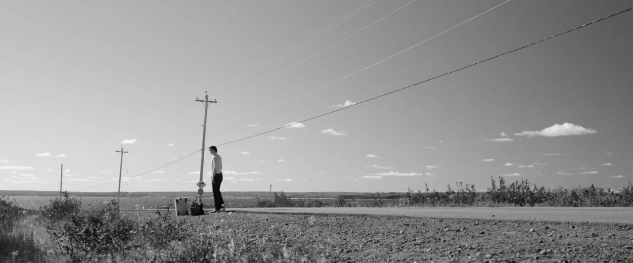 Weirdos (2016) movie still: wide — A long stretch of remote road borders a body of water under a clear sky. A lone person stands near utility poles…